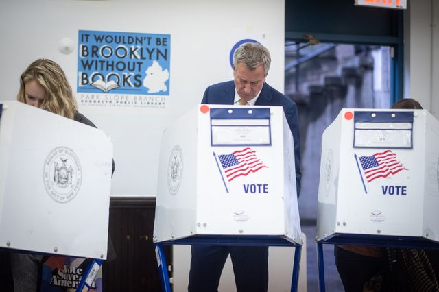 With his head down, Mayor de Blasio stands behind a voting booth stand that includes an image of an American flag and the word vote on it.